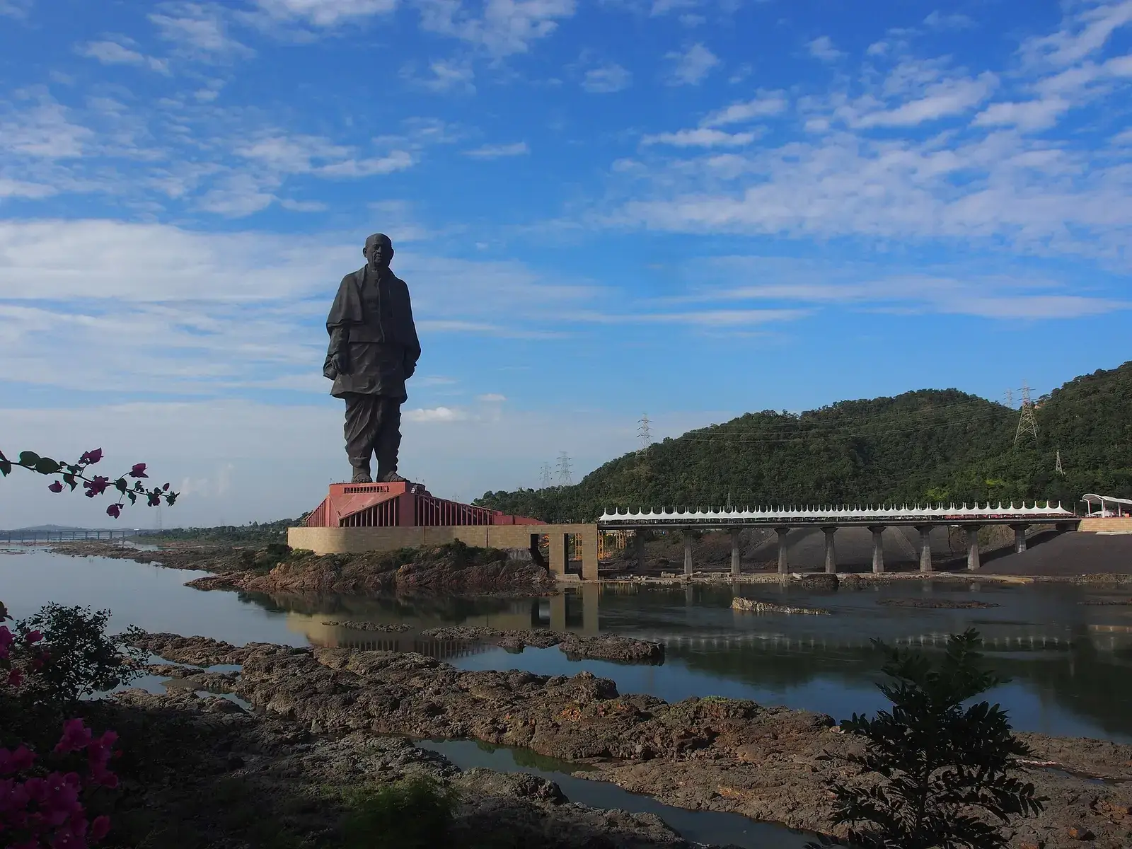 The Statue of Unity seen across the Narmada, with green hills behind