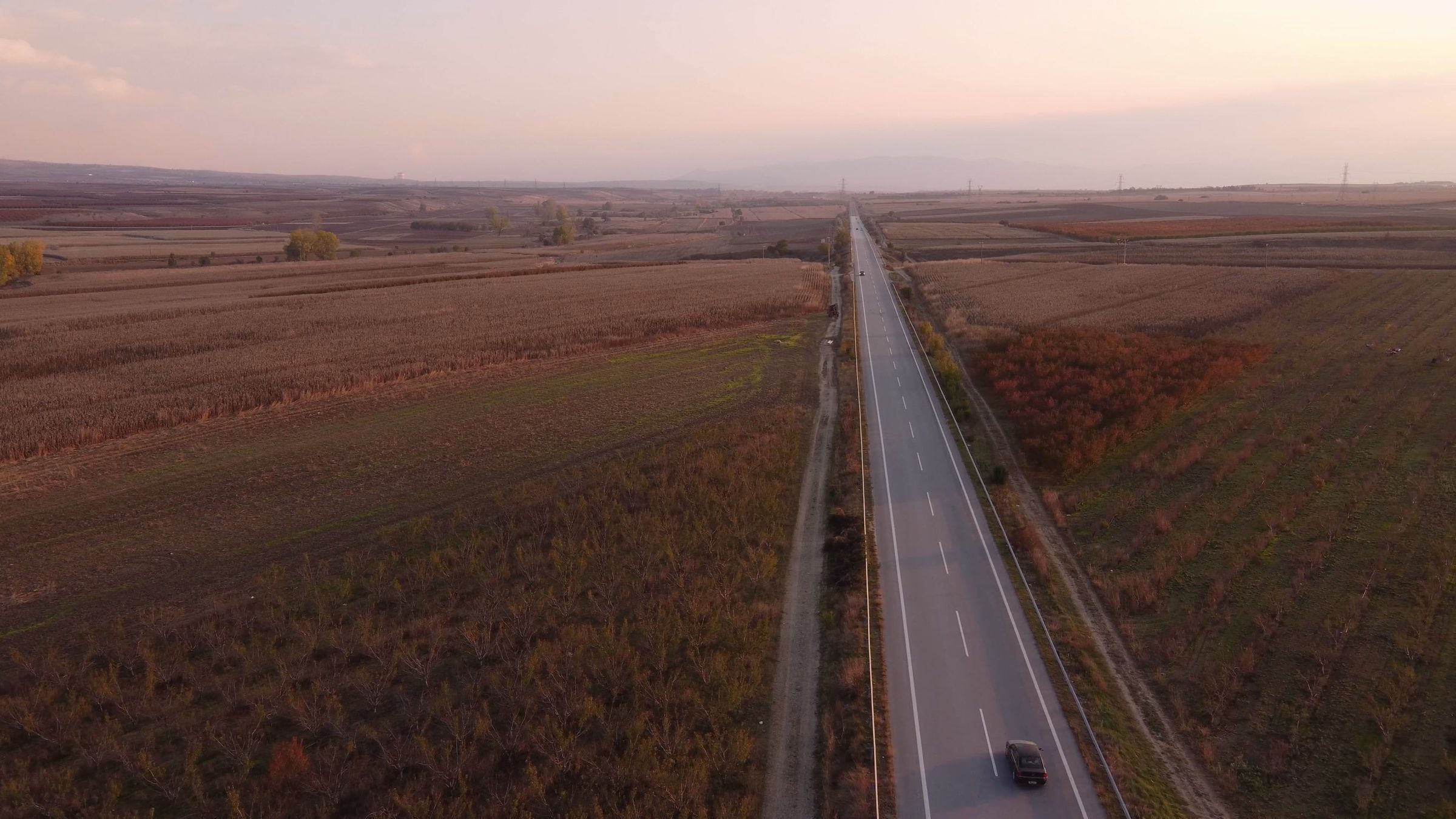 An empty highway through autumn fields at dusk, a single car driving it